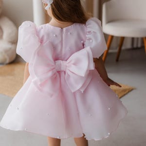 Puede incluir: Un vestido de tul rosa con un gran lazo rosa en la espalda. El vestido est&aacute; decorado con peque&ntilde;as perlas blancas.