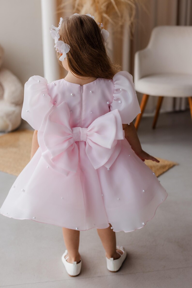 Puede incluir: Un vestido de tul rosa con un gran lazo rosa en la espalda. El vestido est&aacute; decorado con peque&ntilde;as perlas blancas.