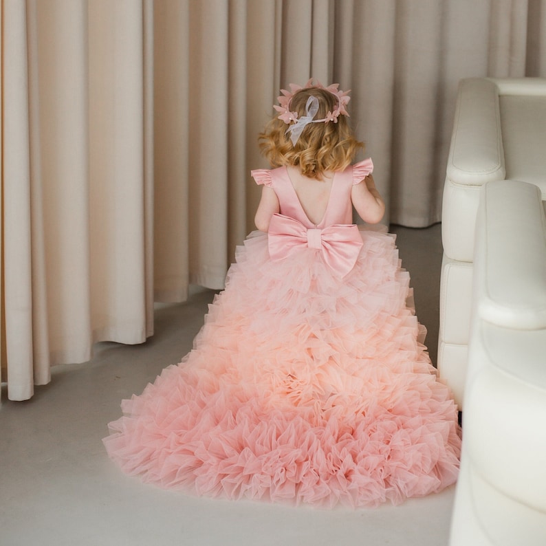 May include: A young girl wearing a pink tulle dress with a large pink bow on the back. The dress has a gradient of pink shades, starting with a darker pink at the top and fading to a lighter pink at the bottom. The dress is very full and has many layers of tulle.