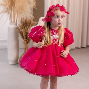 Puede incluir: Una ni&ntilde;a peque&ntilde;a con un vestido rosa con adornos de perlas y una diadema de flores rosas. El vestido tiene mangas cortas abullonadas y una falda completa.