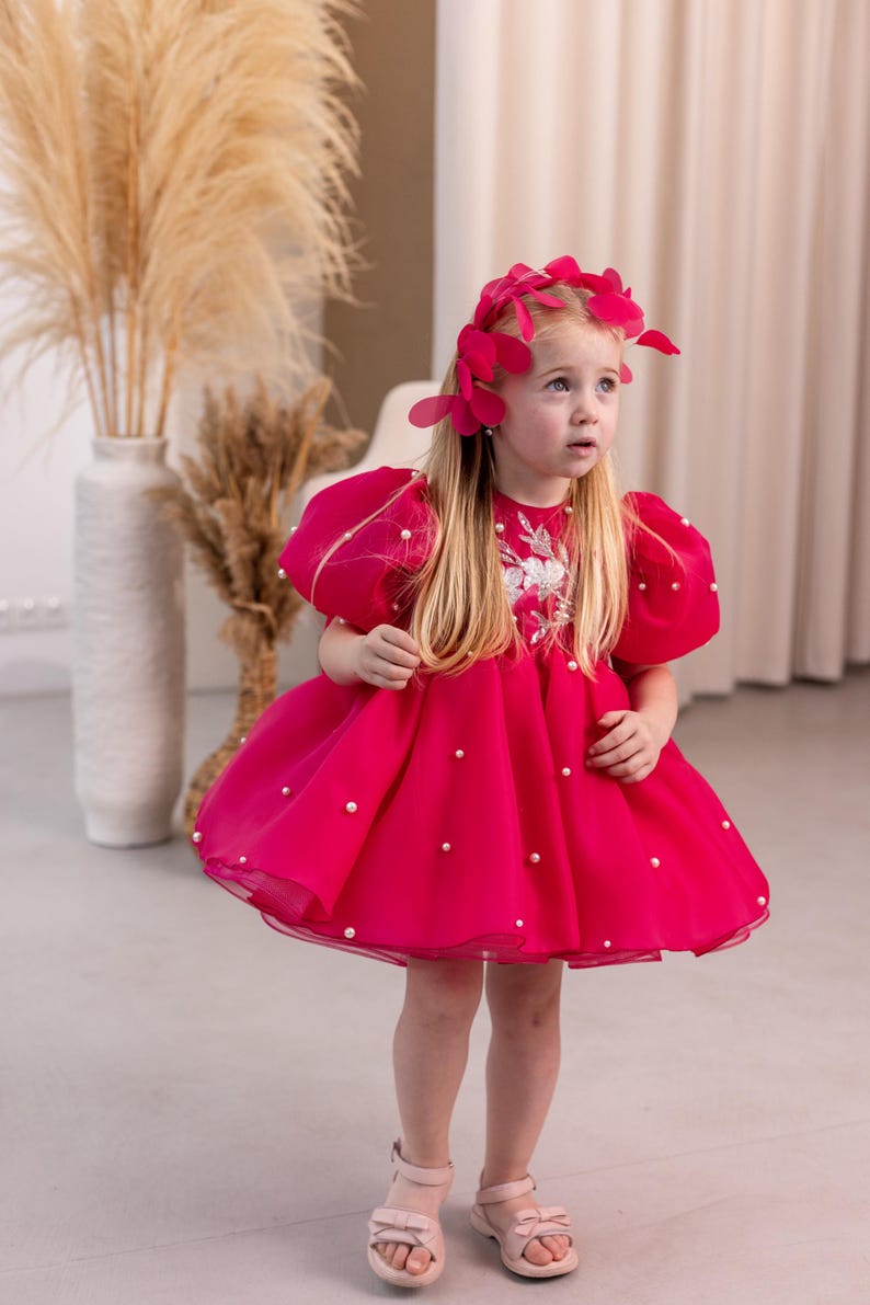 Puede incluir: Una ni&ntilde;a peque&ntilde;a con un vestido rosa con adornos de perlas y una diadema de flores rosas. El vestido tiene mangas cortas abullonadas y una falda completa.