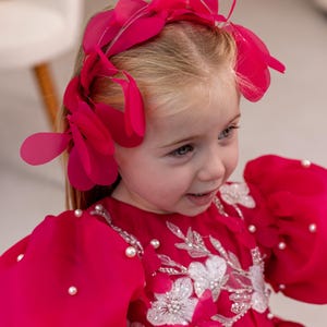 Puede incluir: Una ni&ntilde;a peque&ntilde;a con un vestido rosa brillante con adornos de perlas y bordados florales blancos. Lleva una diadema de flores rosas.