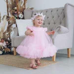 Peut inclure: Une jeune fille portant une robe en tulle rose avec un bandeau papillon. Elle est debout devant un fauteuil gris et sourit.