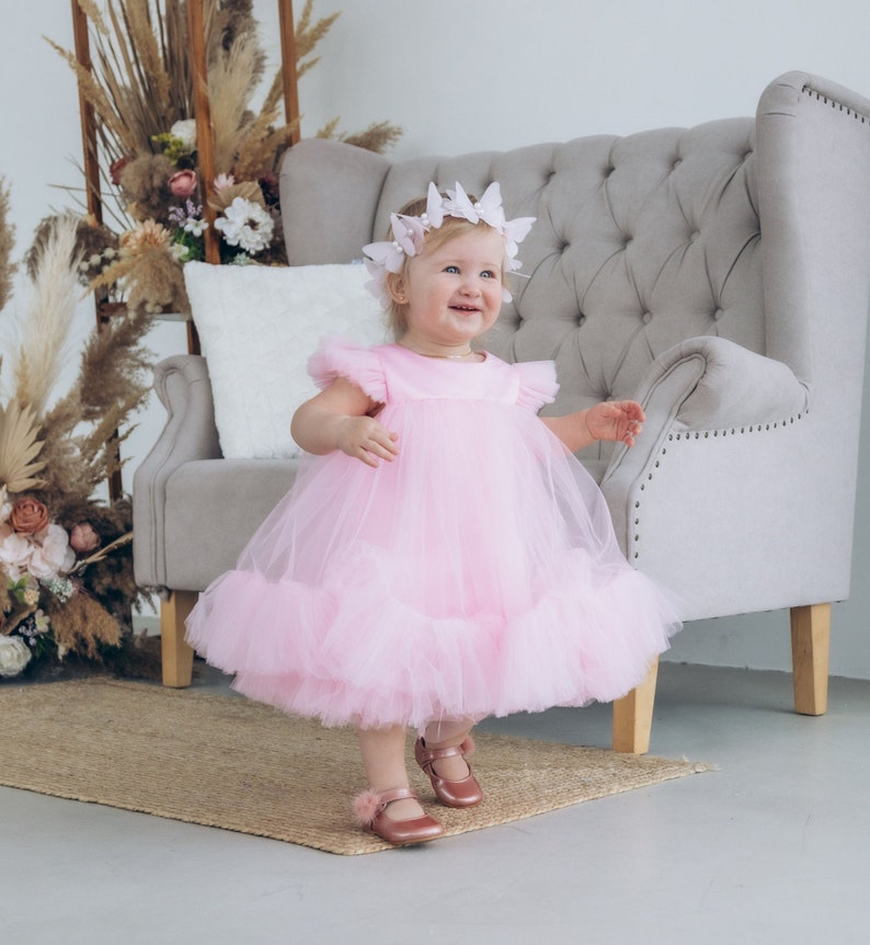 Peut inclure: Une jeune fille portant une robe en tulle rose avec un bandeau papillon. Elle est debout devant un fauteuil gris et sourit.