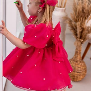 Puede incluir: Una ni&ntilde;a peque&ntilde;a con un vestido rosa con adornos de perlas y una diadema de plumas rosas. El vestido tiene un gran lazo en la espalda.