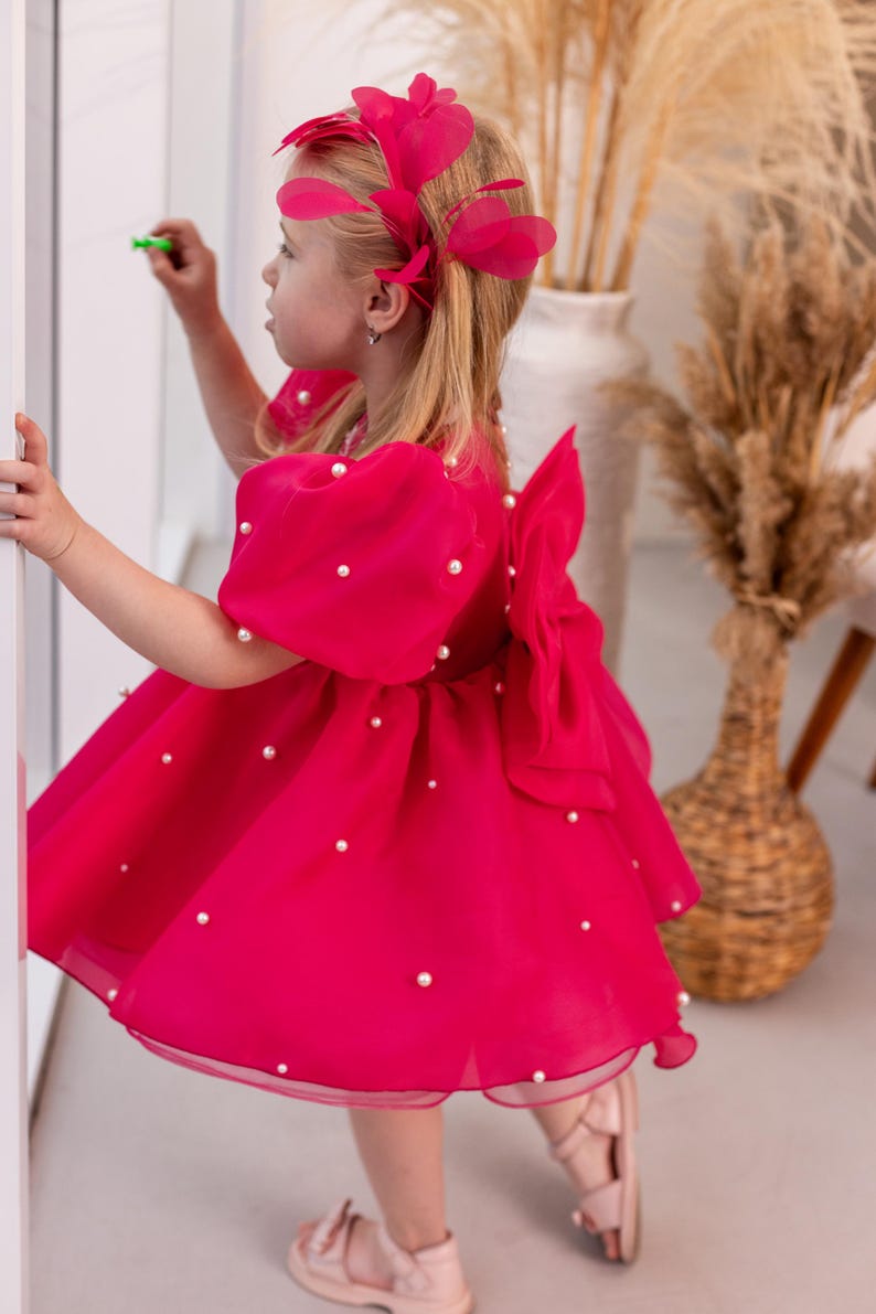 Puede incluir: Una ni&ntilde;a peque&ntilde;a con un vestido rosa con adornos de perlas y una diadema de plumas rosas. El vestido tiene un gran lazo en la espalda.