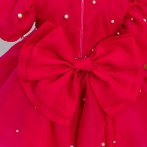 Puede incluir: Un vestido rosa brillante con un gran lazo en la espalda. El vestido est&aacute; decorado con peque&ntilde;as perlas blancas.