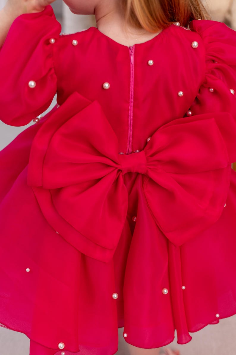 Puede incluir: Un vestido rosa brillante con un gran lazo en la espalda. El vestido est&aacute; decorado con peque&ntilde;as perlas blancas.