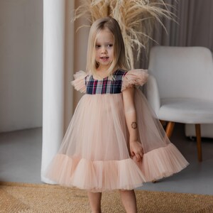 Peut inclure: Une jeune fille portant une robe en tulle rose avec un corsage &agrave; carreaux. La robe a un ourlet &agrave; volants et la fille porte des chaussures blanches.