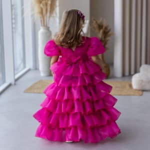Puede incluir: Una niña pequeña con un vestido rosa brillante con múltiples capas de volantes. El vestido tiene un lazo en la espalda y mangas cortas abullonadas.