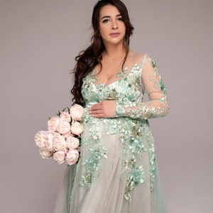 Op de afbeelding: Een zwangere vrouw in een lichtgroene, met bloemen geborduurde jurk met lange mouwen. Ze houdt een boeket witte pioenen vast.