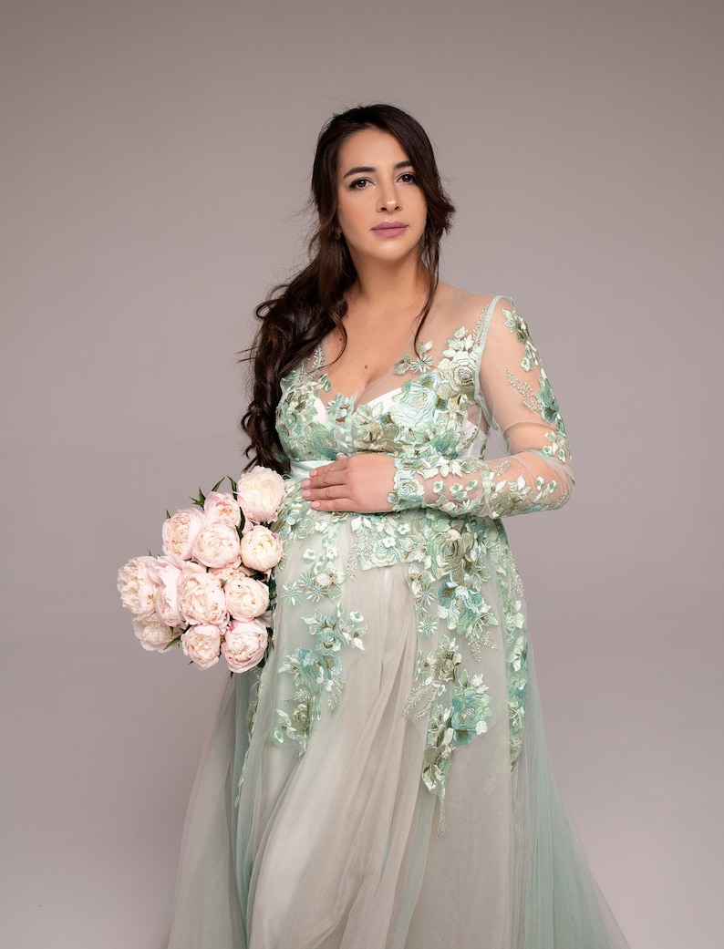 Op de afbeelding: Een zwangere vrouw in een lichtgroene, met bloemen geborduurde jurk met lange mouwen. Ze houdt een boeket witte pioenen vast.