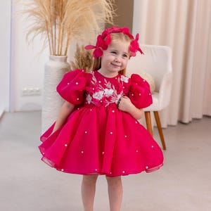Puede incluir: Una ni&ntilde;a peque&ntilde;a con un vestido rosa brillante con adornos de perlas y una diadema de flores rosas. El vestido tiene mangas abullonadas y una falda completa.