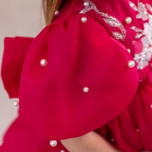 Puede incluir: Un vestido rosa brillante con mangas abullonadas y adornos de perlas. El vestido tiene un dise&ntilde;o de encaje blanco en el corpi&ntilde;o.