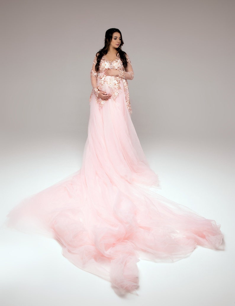 Op de afbeelding: Een zwangere persoon draagt een lange, vloeiende roze tule jurk met delicate kantdetails. De jurk heeft een aansluitend lijfje en een volle rok die achter haar aansleept.