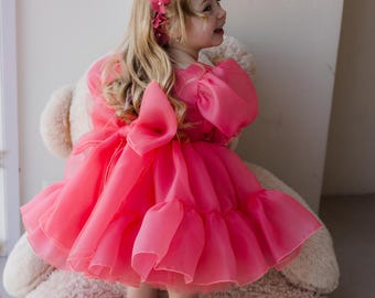 Puede incluir: Una niña pequeña con un vestido de tul rosa con un gran lazo en la espalda. Lleva una corona de flores y sonríe. Está de pie frente a un gran oso de peluche.