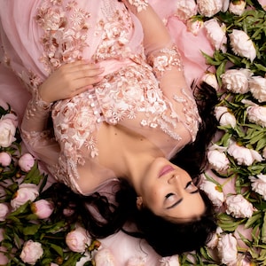 Op de afbeelding: Een zwangere vrouw in een roze, doorschijnende, kanten jurk met bloemenborduursel ligt op een bed van witte pioenrozen. Ze heeft lang donker haar en draagt lichte make-up.