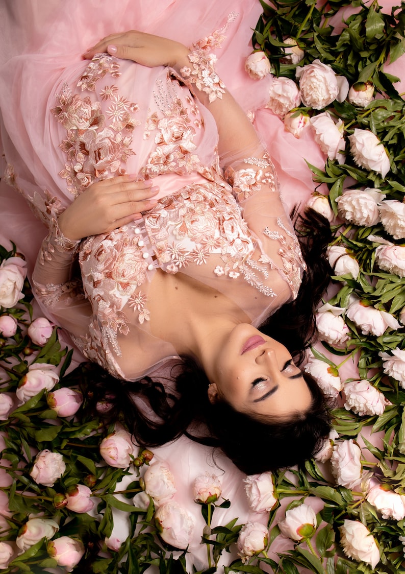 Op de afbeelding: Een zwangere vrouw in een roze, doorschijnende, kanten jurk met bloemenborduursel ligt op een bed van witte pioenrozen. Ze heeft lang donker haar en draagt lichte make-up.