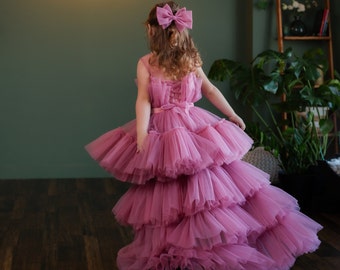 Op de afbeelding: Een jong meisje draagt een roze tulejurk met meerdere lagen. De jurk heeft een aansluitend lijfje en een volle rok. Het meisje draagt een roze strik in haar haar.
