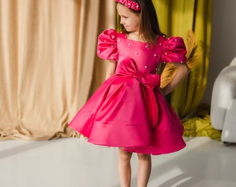 Puede incluir: Una niña pequeña con un vestido de satén rosa con adornos de perlas y un lazo rosa. Lleva una diadema rosa y zapatos negros con un logotipo blanco.