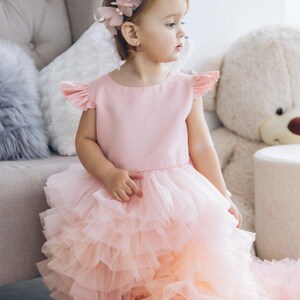 May include: A young girl wearing a pink dress with a tulle skirt and a flower crown. The dress has a pink bodice and a layered tulle skirt. The girl is sitting on a chair and looking to the side.