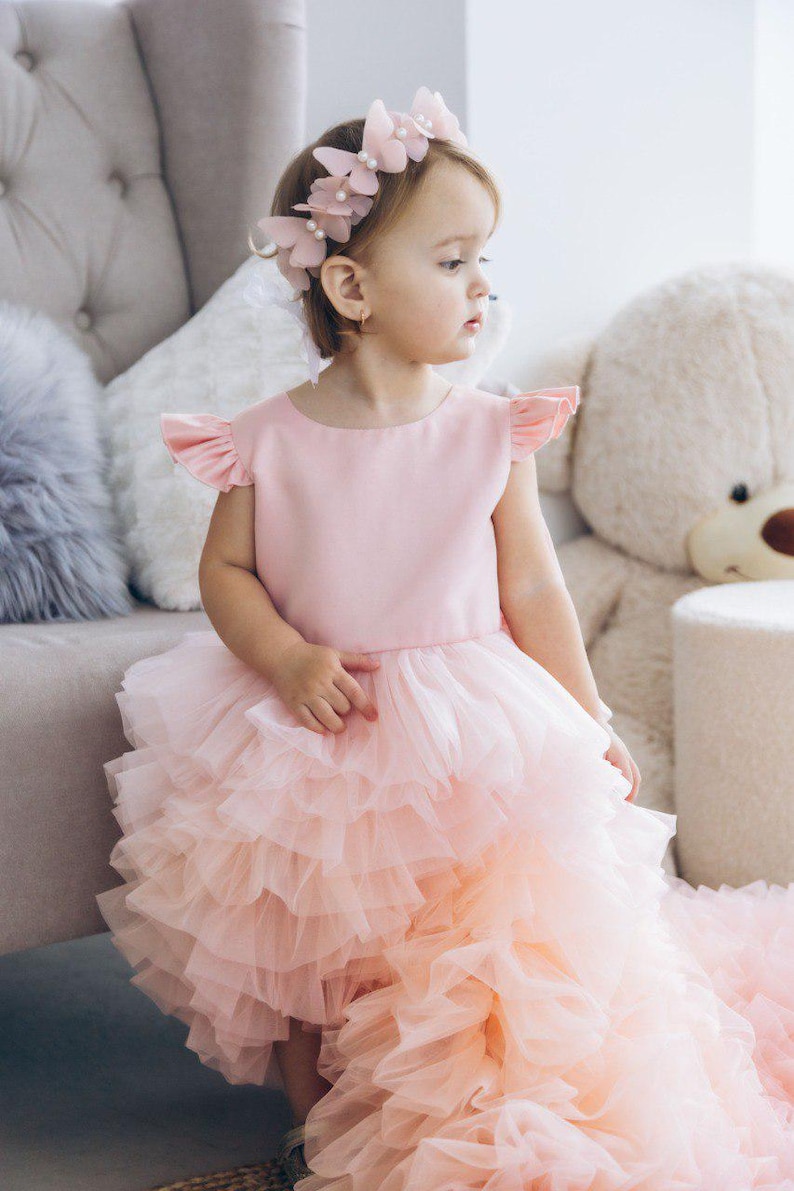 May include: A young girl wearing a pink dress with a tulle skirt and a flower crown. The dress has a pink bodice and a layered tulle skirt. The girl is sitting on a chair and looking to the side.