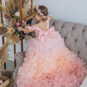 May include: A young girl wearing a pink and peach ombre tulle dress with a large pink bow on the back. The dress has multiple layers of tulle and is very full. The girl is sitting on a gray couch and is looking down at something.