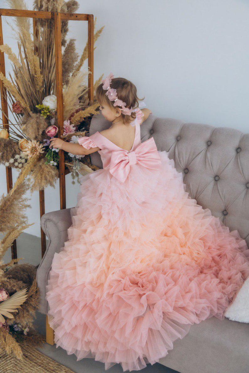 May include: A young girl wearing a pink and peach ombre tulle dress with a large pink bow on the back. The dress has multiple layers of tulle and is very full. The girl is sitting on a gray couch and is looking down at something.