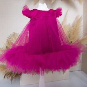 Peut inclure: Une robe en tulle magenta avec un corsage en satin et des manches &agrave; volants. La robe est sur un mannequin blanc.