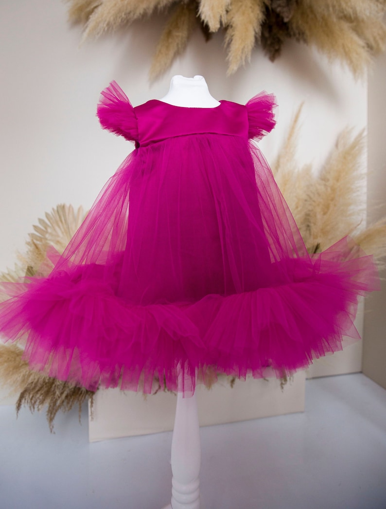 Peut inclure: Une robe en tulle magenta avec un corsage en satin et des manches &agrave; volants. La robe est sur un mannequin blanc.