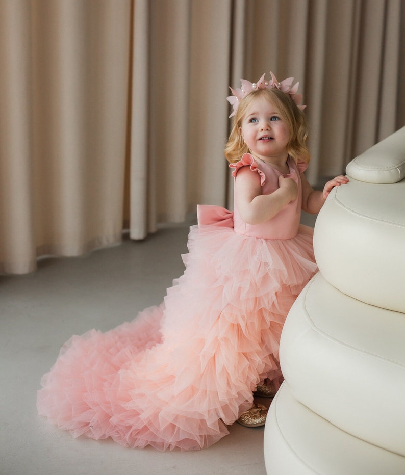 May include: A young girl wearing a pink tulle dress with a long train and a flower crown. She is standing next to a white couch.