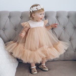 Peut inclure: Une jeune fille portant une robe en tulle p&ecirc;che avec une couronne de fleurs blanches et des chaussures dor&eacute;es &agrave; paillettes. Elle est assise sur un canap&eacute; gris.