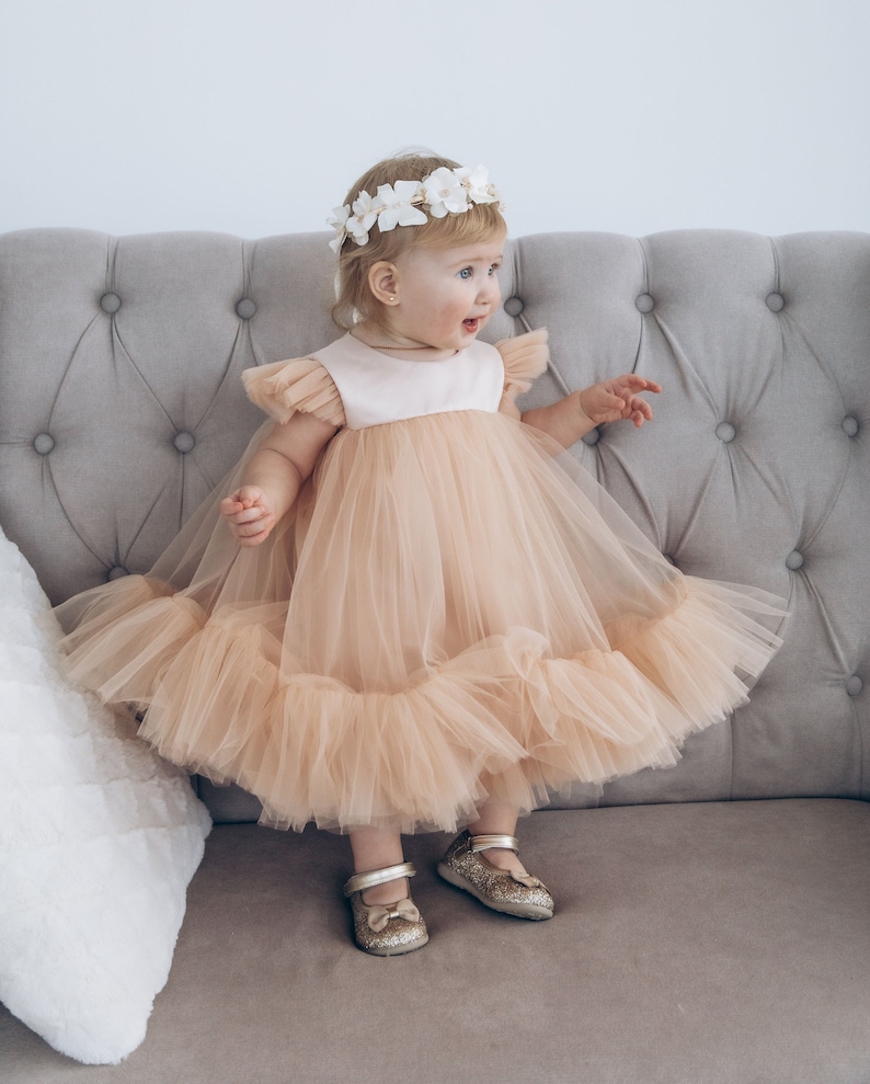 Peut inclure: Une jeune fille portant une robe en tulle p&ecirc;che avec une couronne de fleurs blanches et des chaussures dor&eacute;es &agrave; paillettes. Elle est assise sur un canap&eacute; gris.