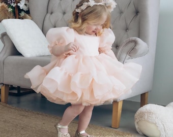 Puede incluir: Una niña pequeña con un vestido de tul rosa claro con falda con volantes y mangas abullonadas. Lleva una corona de flores y zapatos dorados. La niña está de pie frente a un sillón gris.