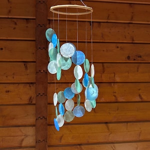 Peut inclure: Un carillon éolien suspendu, composé d'un anneau en bois et de fils supportant des disques circulaires irisés bleus, verts et blancs. Les disques de tailles variées sont suspendus devant un fond de planches de bois.