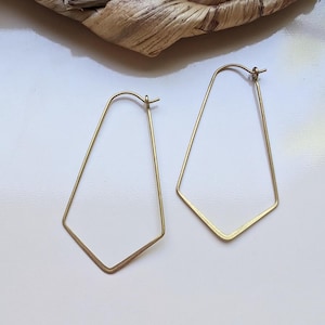 Könnte beinhalten: Ein Paar goldfarbene geometrische Creolen. Die Ohrringe haben eine einzigartige, eckige Form mit flachem Boden und spitzer Spitze. Die Ohrringe werden auf einer weißen Oberfläche präsentiert.