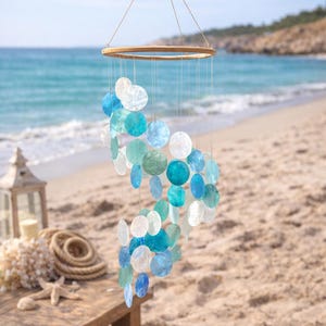 Puede incluir: Un carillón de viento colgante con un anillo de madera y hebras de acentos circulares azules, turquesas y blancos. El carillón de viento cuelga contra un fondo de playa y océano.