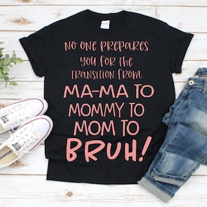 Könnte beinhalten: Schwarzes T-Shirt mit dem Text "No one prepares you for the transition from MA-MA TO MOMMY TO MOM TO BRUH!" in rosa Schrift.