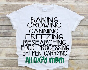 May include: White t-shirt with black text that reads "Baking, Growing, Canning, Freezing, Researching, Food Processing, Epi Pen Carrying, Allergy Mom". The word "Mom" is in green.