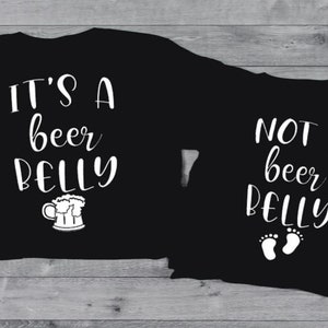 Può includere: Due magliette nere con testo bianco. La maglietta più grande dice "It's a beer belly" con un'immagine di una boccale di birra. La maglietta più piccola dice "Not a beer belly" con due impronte di piedi.