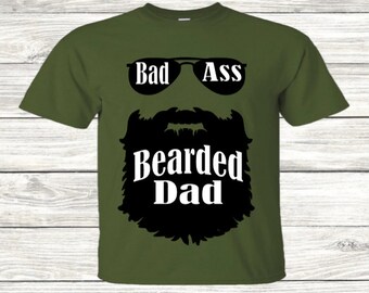 Pode incluir: Uma camiseta verde com uma silhueta preta de barba e óculos de sol. O texto "Bad Ass Bearded Dad" está impresso em branco na camiseta.