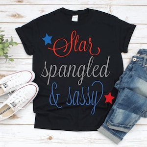 May include: A black t-shirt with the text "Star spangled & sassy" in red, blue, and gray lettering. There are red and blue stars on either side of the text. The shirt is laid out on a white wooden surface with a pair of white sneakers and blue jeans.