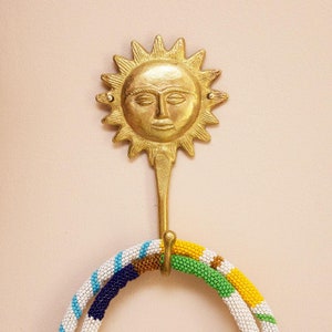 Könnte beinhalten: Goldener Haken in Sonnenform mit einem Gesicht. Eine bunte Perlenkette hängt an dem Haken.