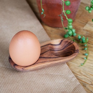 May include: A brown egg sits in a wooden egg holder. The holder is made of dark wood with a natural grain pattern. The egg holder is on a brown paper surface.