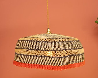 Puede incluir: Una lámpara colgante en forma de cúpula hecha de fibras naturales tejidas, con bandas horizontales alternas en color beige y marrón oscuro, terminada con un borde naranja vibrante en la base y suspendida por un cable beige a juego.