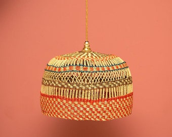 Puede incluir: Una lámpara colgante tejida con una combinación de colores natural, beige y rojo. La lámpara tiene una forma redondeada, similar a una campana, con un patrón tejido. La luz cuelga de un accesorio y un cable dorados, creando un ambiente cálido.