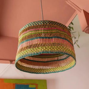 Op de afbeelding: Een geweven hanglamp met een cilindrische vorm. De lamp heeft horizontale strepen in groen, roze en bruin. Hij hangt aan een zwarte plafondbevestiging.
