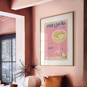 Peut inclure: Une affiche encadr&eacute;e rose et or avec le mot "Margarita" en bleu. L'affiche repr&eacute;sente un cocktail margarita rose avec un quartier de citron vert et un motif d'&eacute;toiles. L'affiche est accroch&eacute;e &agrave; un mur rose dans une salle &agrave; manger avec une table ronde et des chaises.