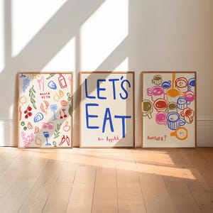 May include: Three framed art prints leaning against a wall. The prints feature colourful illustrations of food, drinks, and the words "LET'S EAT" and "ANOTHER?" in bold text. The frames are a light brown colour.