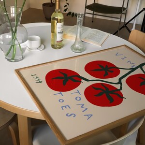 May include: A framed print with a white background and a stylised illustration of red tomatoes on a vine. The text "TOMA TOES" is written in blue on the print. The print is framed in a natural wood frame.